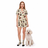 A woman modeling a matching short-sleeve button-front shirt and shorts set with socks, standing next to a small white poodle on a plain white studio background. The shirt and shorts have a comic book vintage-style illustrated print on a light cream base, and the socks match the print.