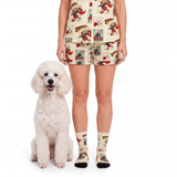 A person wearing a matching beige pajama set with a colorful cartoon dog print sits next to a small white poodle on a white studio background. The set includes a short-sleeve top and shorts, paired with matching patterned crew socks.