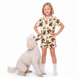 A young girl stands next to a white poodle on a white studio background, wearing a matching short-sleeve pajama set with a comic book vintage-style dog illustration print. The pajamas have a cream base with supergirl prints