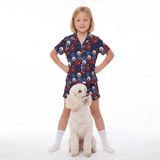 A young girl stands with hands on hips wearing a Supergirl-themed pajama set with white dog print, paired with white socks, next to a small white poodle on a seamless white background.
