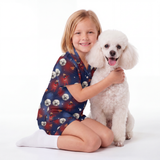 A young girl with blonde hair sits on a white studio floor, hugging a white poodle. She is wearing a short-sleeve Supergirl-themed pajama set with a dog print and white socks.