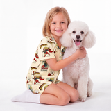 A young girl with blonde hair sits on the floor wearing a cream-colored pajama set with a Supergirl cartoon print, hugging a white poodle dog against a plain white background.