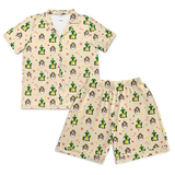A two-piece pajama set featuring a short-sleeve button-up shirt and matching shorts in a warm cream color, both printed with a whimsical pattern of green cacti, brown and white dogs, red candy canes, and small yellow stars.