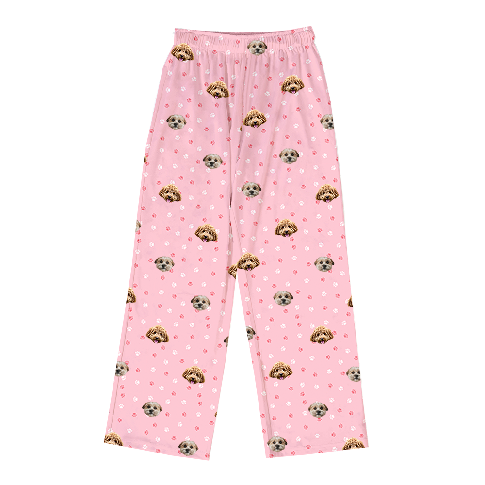 A pair of full-length pajama pants in light pink. The pants feature a repeating pattern of cartoonish pink and white paw prints, interspersed with realistic photos of two different dogs' heads.