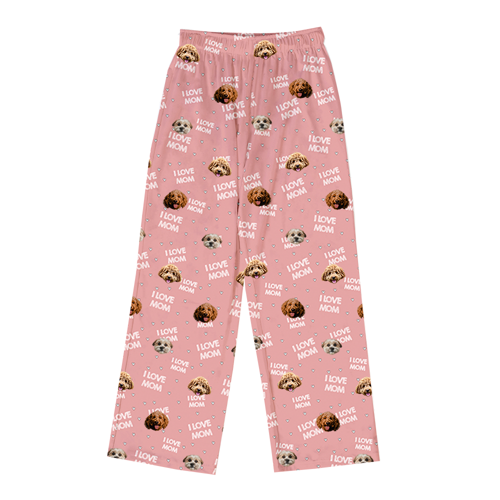A pair of pink full-length pajama pants featuring a repeating pattern of various dog faces in different shades of brown and white, and white text reading "I LOVE MOM" interspersed with small white hearts.