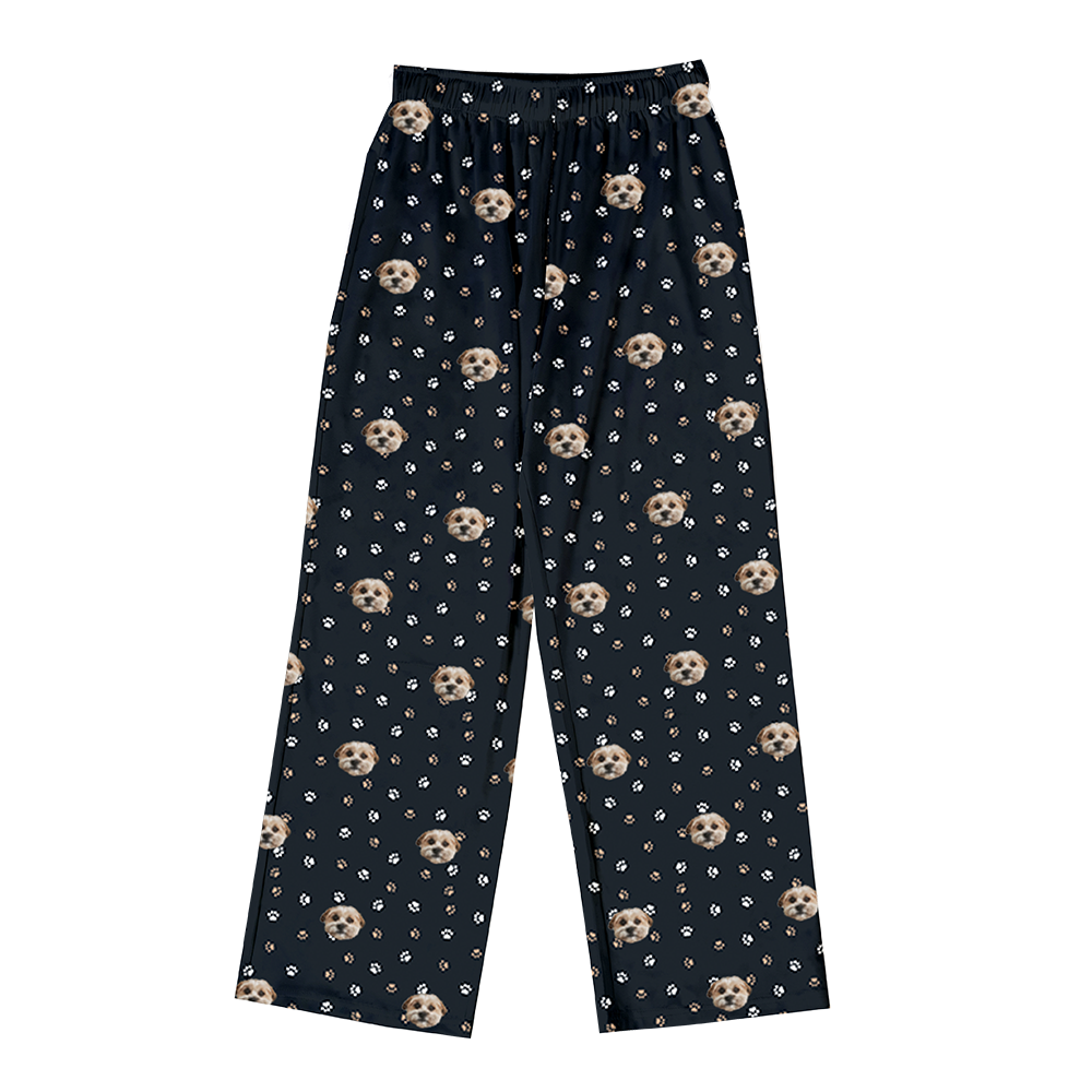 A pair of black full-length pajama pants featuring a repeating pattern of light brown and white dog faces interspersed with scattered white and light brown paw prints. 