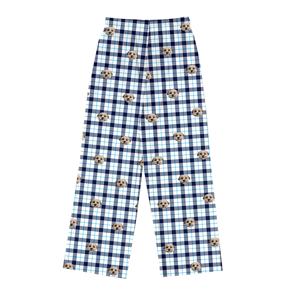 A pair of full-length pajama pants featuring a white background with a large navy blue and thin light blue plaid pattern. Interspersed across the plaid is a repeating pattern of light brown and white dog faces.