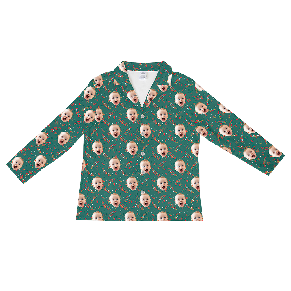 Long-sleeved, button-down pajama shirt in dark teal green with a repeating pattern of a cheerful baby's face, small white dots, and sprigs of red berries and pine needles, suggesting a holiday theme.