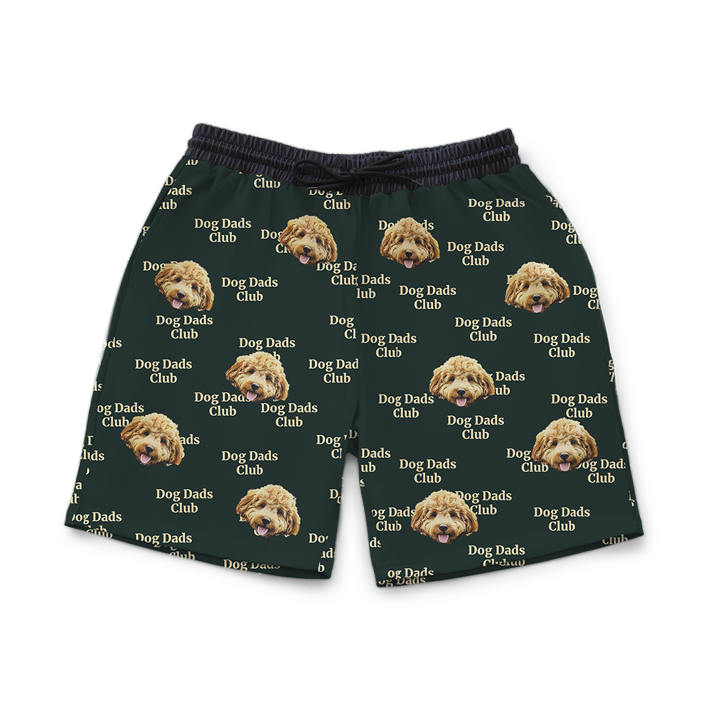Green athletic shorts featuring a repeating pattern of a fluffy light-brown dog’s face and the phrase “Dog Dads Club” in beige text.
