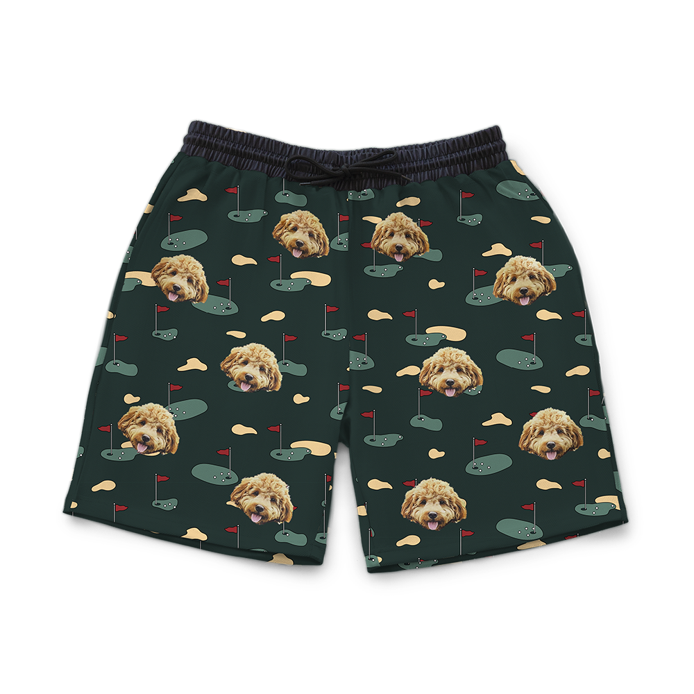 Green men’s shorts printed with a repeating golf-course pattern featuring small putting greens, sand traps, and red flags, decorated with multiple faces of a fluffy tan dog.
