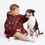 A young boy wearing a burgundy hooded sweatshirt with a dog print sits on a white studio floor and pets a black, white, and tan dog.
