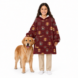 A young girl stands next to a golden retriever dog wearing a burgundy long-sleeve hoodie with a repeating print of golden retriever dog faces and small decorative motifs. The girl is also wearing beige pants and white socks against a plain white background.