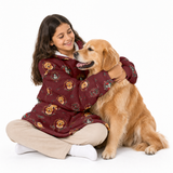 A young girl with long dark hair sits cross-legged on a white studio floor, hugging a golden retriever dog. She is wearing a burgundy fleece pajama hoodie with a playful print of cartoon dogs, cats, and birds, paired with beige pants and white socks.