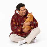A man sitting cross-legged on a white studio background, holding an orange tabby cat. He is wearing a plush hooded blanket sweater in a deep burgundy red with a colorful cartoon cat pattern.