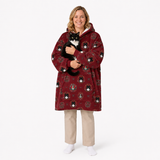 A woman wearing a red hooded blanket sweater with a black and white cat wrapped in it, standing against a plain white studio background.