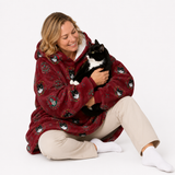 A woman sitting on a white studio floor holds a black and white cat wrapped in a plush hooded blanket. The blanket is burgundy red with a repeating pattern of cartoon cats' faces and small decorative motifs.
