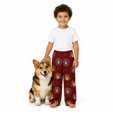 A young boy stands next to a corgi dog on a white studio background, wearing a white t-shirt and burgundy pajama pants with a repeating crest pattern. The boy is smiling and looking at the camera, with the dog sitting beside him.