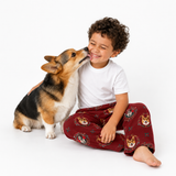 A child wearing red pajama pants with a corgi dog print sits on a white studio floor, smiling as a corgi licks their face. The child is also wearing a plain white t-shirt.