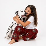 A young girl with long brown hair sits on the floor hugging a Dalmatian dog against a plain white studio background. She is wearing a white short-sleeve T-shirt and red pajama pants printed with Dalmatian faces and decorative frames.