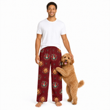 A man wearing burgundy pajama pants with a gold and green dog pattern stands barefoot next to a small golden dog on a white background.