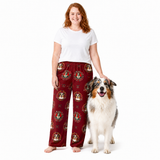 A woman stands barefoot next to a dog wearing red pajama pants with a repeating dog-face pattern, against a plain white studio background.