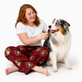 A woman with red hair sits on the floor brushing a tricolor Australian Shepherd dog. She is wearing a white t-shirt and red pajama pants with a dog face pattern.