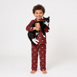 A young boy stands barefoot against a light gray studio background, smiling and holding a black-and-white cat. He is wearing a matching red pajama set with a small repeating festive print.