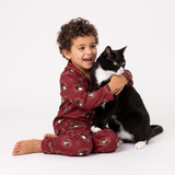 A young child with curly brown hair wearing a burgundy pajama set with a cartoon penguin print sits on a white studio floor, hugging a black and white cat.