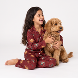 A young girl with long dark hair sits on a white studio floor hugging a fluffy golden-brown dog. She is wearing a matching burgundy pajama set with a playful owl pattern, and the photo is lit with bright, even studio light.
