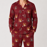 A man wearing a matching long-sleeve button-up pajama shirt and pants set in deep burgundy with an all-over print of corgi dog faces and ornate crest motifs, photographed against a light gray studio background.