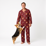 A man wearing a burgundy long-sleeve pajama set with a repeating fox print stands barefoot against a white studio background, with a corgi dog standing on its hind legs and placing its paw on his thigh.