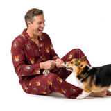 A man wearing a maroon pajama set with a dog print sits on a white studio floor, smiling and petting a corgi dog.