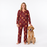 A woman wearing a burgundy long-sleeve pajama set with a repeating dog face pattern stands barefoot on a white studio background, smiling and petting a golden retriever dog.