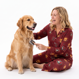 A woman in a maroon pajama set with a cartoon lion pattern sits on a white studio floor, smiling as she brushes a golden retriever dog.
