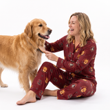 A woman wearing a maroon pajama set with a pumpkin print sits on the floor next to a golden retriever dog, smiling and petting it, against a solid white background.