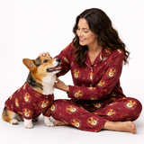 A woman and a corgi dog wearing matching burgundy pajamas with a dog-themed print, sitting together on a white studio background.