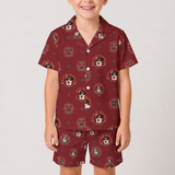 A child wearing a matching short-sleeve button-front pajama shirt and shorts set in burgundy with a repeating printed pattern of dog faces and decorative motifs, photographed against a light gray studio background.