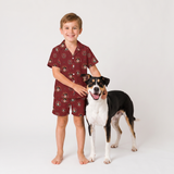 A young boy wearing a matching short-sleeve button-up pajama shirt and shorts set with a dog print, standing next to a black, white, and tan dog on a seamless white studio background.