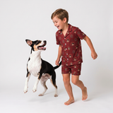 A young boy wearing a short-sleeve pajama set with a red-burgundy base and a cartoon monkey print, running playfully next to a black and white dog on a seamless white studio background.
