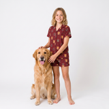 A young girl stands barefoot next to a golden retriever dog on a white studio background, wearing a matching short-sleeve button-up pajama shirt and shorts set in burgundy with a small multicolor lion pattern.