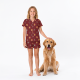 A young girl wearing a short-sleeve button-front pajama shirt and matching shorts with a small crest pattern, standing barefoot next to a golden retriever on a plain white studio background.
