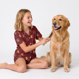 A young girl with blonde hair sits cross-legged on a white studio floor, smiling as she brushes a golden retriever dog. She is wearing a short-sleeve pajama set in maroon with a repeating lion print.
