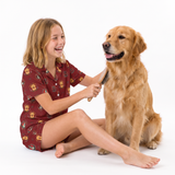 A young girl with blonde hair wearing a red patterned pajama set sits on a white studio floor, brushing a golden retriever dog's fur with a brush.
