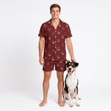 A man wearing a matching short-sleeve button-up pajama shirt and shorts set in a deep red-burgundy color with a small cartoon dog print, standing next to a black, white, and tan dog on a white studio background.