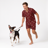 A man wearing a matching short-sleeve button-up pajama shirt and shorts set in a burgundy red color with a cartoon dog print, running barefoot while holding a leash attached to a black, white, and tan dog on a white studio background.