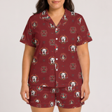 A woman modeling a matching short-sleeve button-front pajama shirt and shorts set in burgundy with a repeating golden retriever and crest pattern, against a light gray studio background.