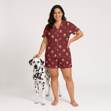 A woman modeling a matching burgundy short-sleeve button-up pajama shirt and shorts set with a white dog print, standing next to a Dalmatian on a light gray studio background.