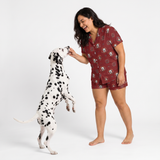 A woman wearing a matching short-sleeve button-up pajama shirt and shorts set with a dog print, interacting playfully with a Dalmatian dog on a white studio background.