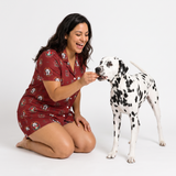 A woman kneels on a white studio floor smiling and offering a treat to a Dalmatian dog, wearing a short-sleeve burgundy pajama set with a dog print. The dog wears a white collar with a black tag.