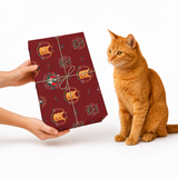 A person's hands hold a wrapped gift box with a dark red patterned wrapping paper next to an orange tabby cat on a white background.